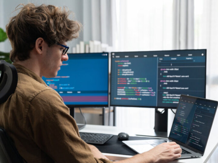 Man working at a desk with multiple monitors displaying code in a bright room.